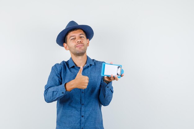 Молодой человек держит буфер обмена с большим пальцем руки вверх в голубой рубашке, шляпе и выглядит весело. передний план.
