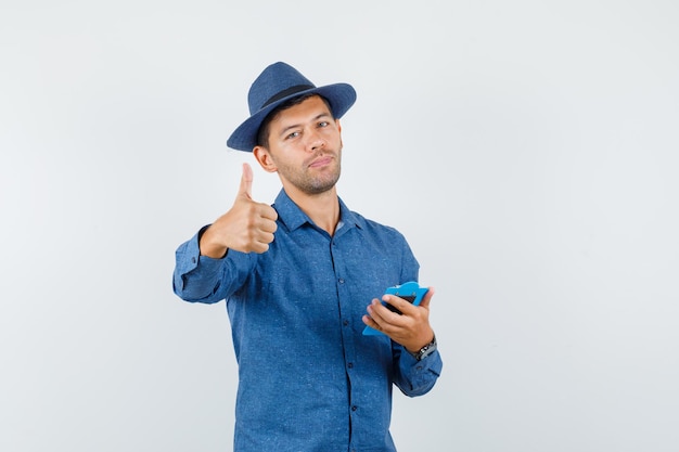 Young man holding clipboard with thumb up in blue shirt, hat and looking cheery. front view.