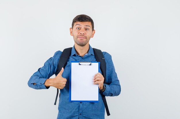 Young man holding clipboard with thumb up in blue shirt and looking pleased. front view.