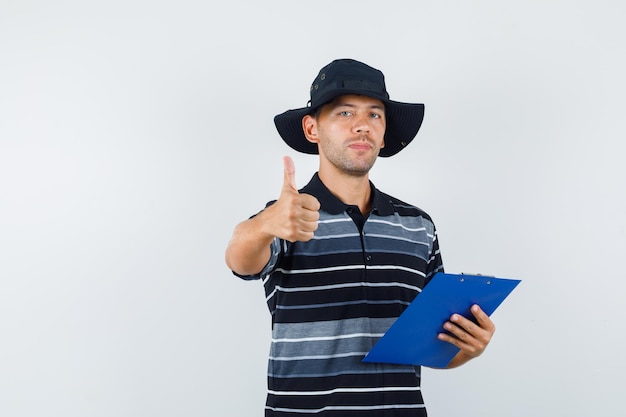 Young man holding clipboard with thumb up in t-shirt, hat and looking pleased. front view.