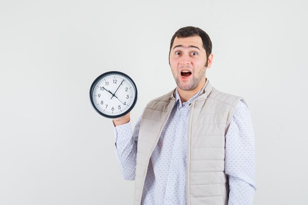 Young man holding clock in one hand in beige jacket and cap and looking surprised. front view.