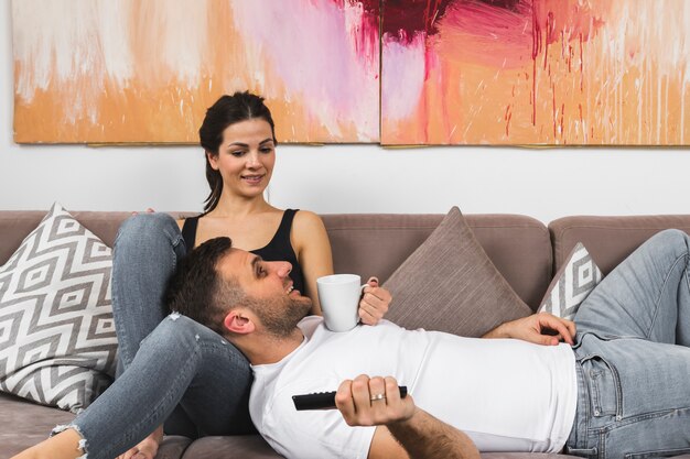 Young man holding coffee cup and remote control lying on woman's lap