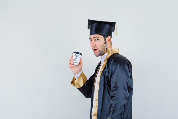 Young man holding cup of coffee in graduate uniform and looking confident. .