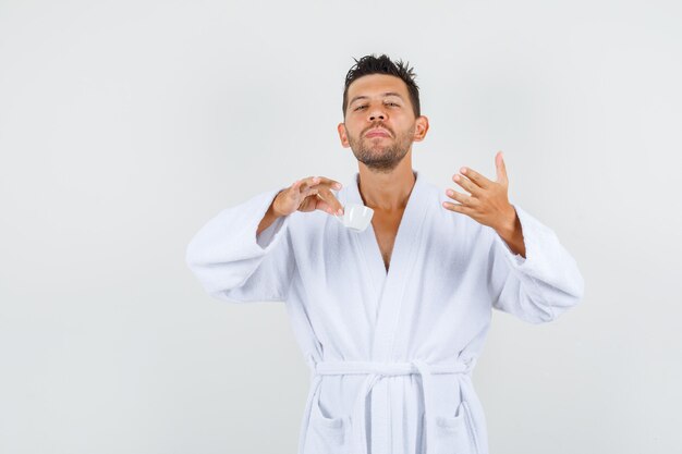 Young man holding cup of coffee in white bathrobe and looking spirited , front view.