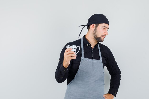 Young man holding cup of drink in shirt, apron and looking concerned , front view.