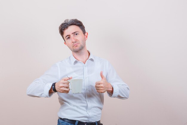 Young man holding cup of drink, showing thumb up in white shirt, jeans