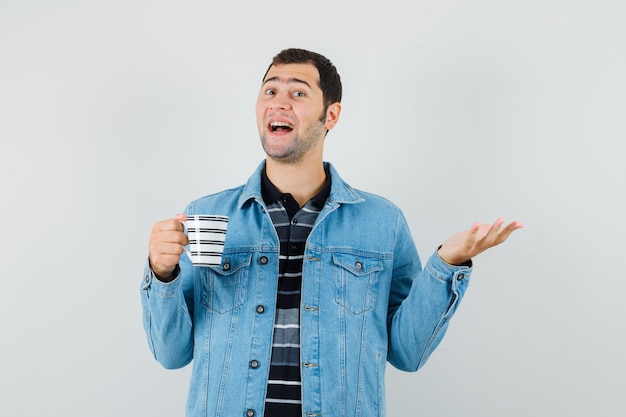 Young man holding cup of drink, spreading palm aside in t-shirt, jacket and looking jovial. 