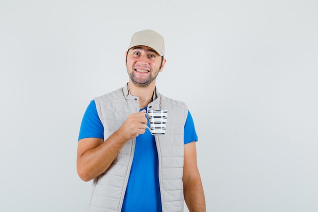 Giovane che tiene tazza di bevanda in t-shirt, giacca e sembra allegro. vista frontale.