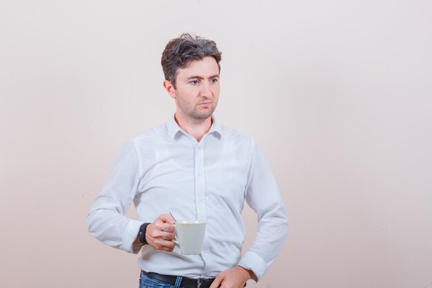 Молодой человек держит чашку напитка в белой рубашке, джинсах и смотрит задумчиво