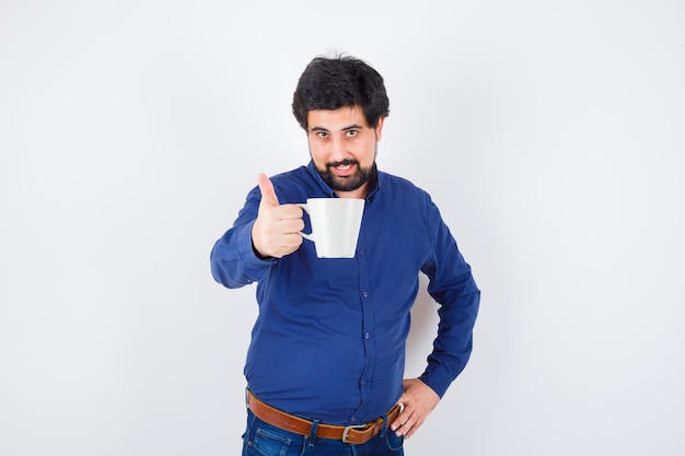 Young man holding cup and holding hand on waist in blue shirt and jeans and looking optimistic. front view.