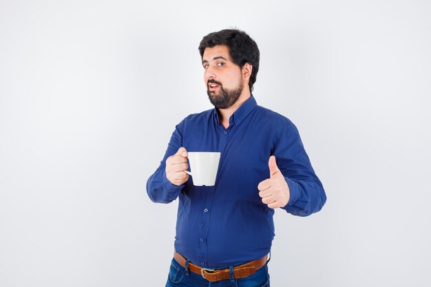 Young man holding cup and showing thumb up in blue shirt and jeans and looking optimistic. front view.