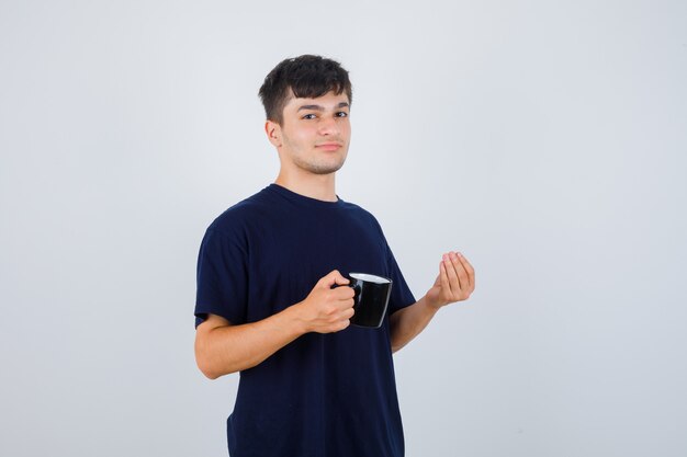 Young man holding cup of tea, doing Italian gesture in black t-shirt and looking delighted. front view.