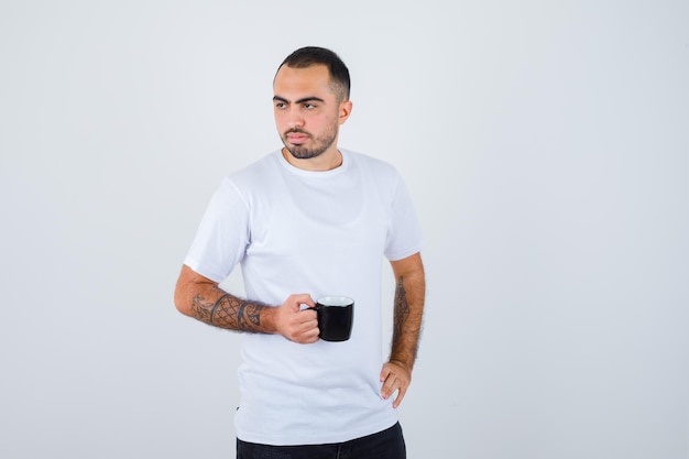 Young man holding cup of tea and holding hand on waist in white t-shirt and black pants and looking serious