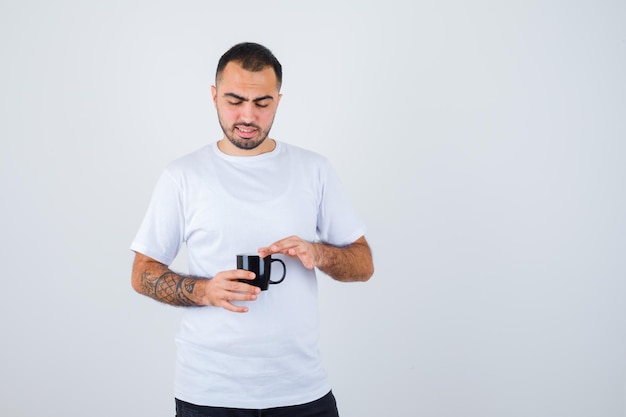 Young man holding cup of tea and putting hand on it in white t-shirt and black pants and looking focused