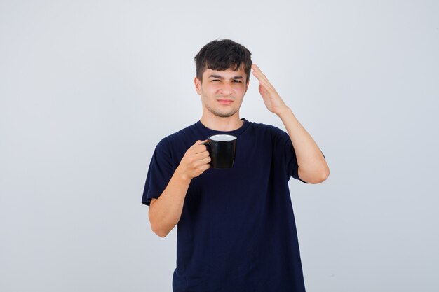 Young man holding cup of tea, raising hand in black t-shirt and looking puzzled , front view.