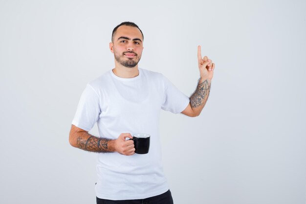 Young man holding cup of tea and raising index finger in eureka gesture in white t-shirt and black pants and looking happy