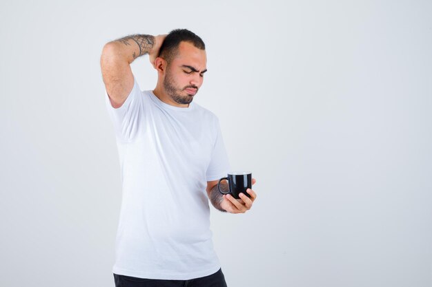 Young man holding cup of tea and scratching head in white t-shirt and black pants and looking serious