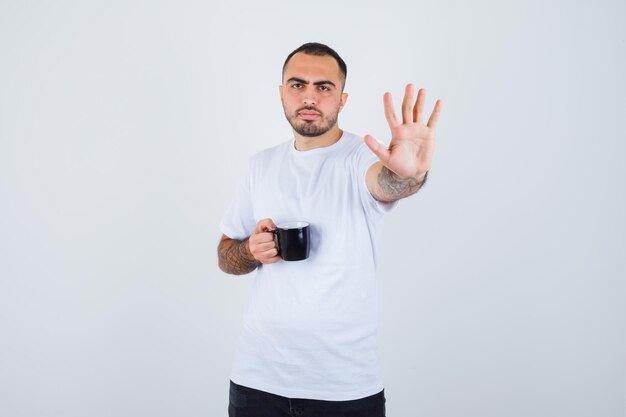 Молодой человек держит чашку чая и показывает стоп в белой футболке и черных штанах и выглядит серьезным