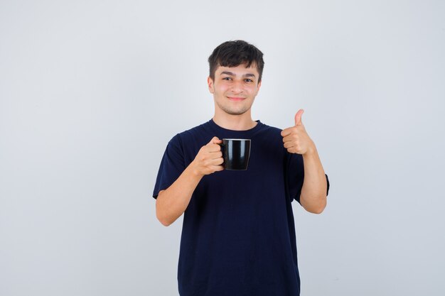 Young man holding cup of tea, showing thumb up in black t-shirt and looking cheery. front view.