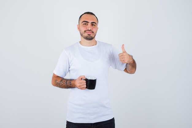 Young man holding cup of tea and showing thumb up in white t-shirt and black pants and looking happy