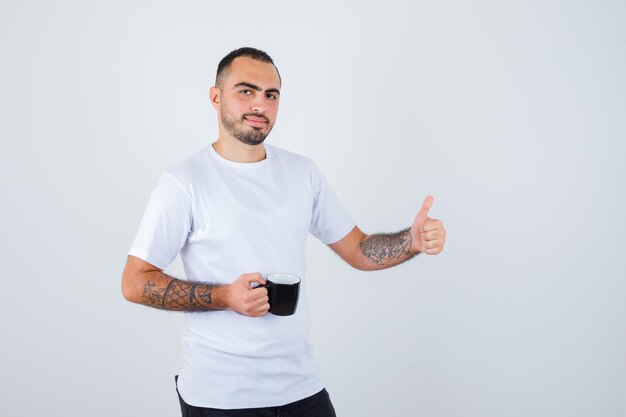 Young man holding cup of tea and showing thumb up in white t-shirt and black pants and looking happy