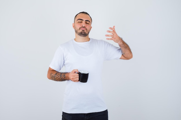 Young man holding cup of tea and stretching hand as holding something imaginary while closing eyes in white t-shirt and black pants and looking calm