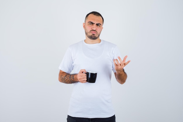 Молодой человек, держащий чашку чая и вопросительно протягивающий руку в белой футболке и черных штанах, выглядит озадаченным