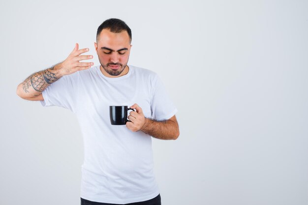 Young man holding cup of tea and stretching hand toward it in white t-shirt and black pants and looking focused