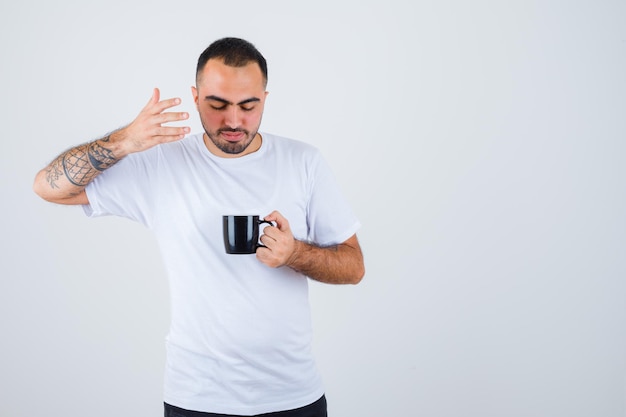 Молодой человек держит чашку чая и протягивает к ней руку в белой футболке и черных штанах и выглядит сосредоточенным