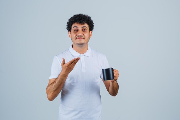 Young man holding cup of tea, stretching hand toward it in white t-shirt and jeans and looking displeased , front view.