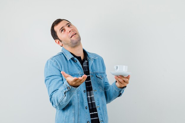 Young man holding cup of tea in t-shirt, jacket and looking confused