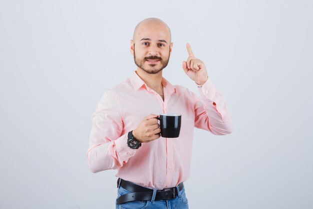 Young man holding cup while pointing up in pink shirt,jeans , front view.