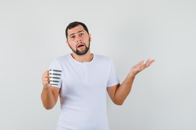 Young man holding cup while showing helpless gesture in white t-shirt and looking confused , front view.