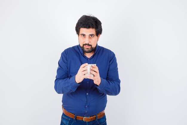 Young man holding cup with both hands in blue shirt and jeans and looking serious. front view.