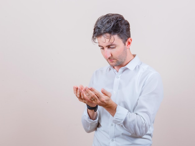 Young man holding cupped hands in praying gesture in white shirt and looking hopeful