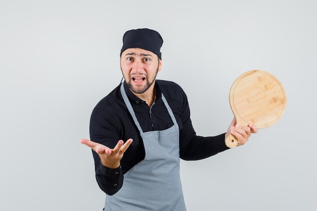 Young man holding cutting board in shirt, apron and looking anxious. front view.