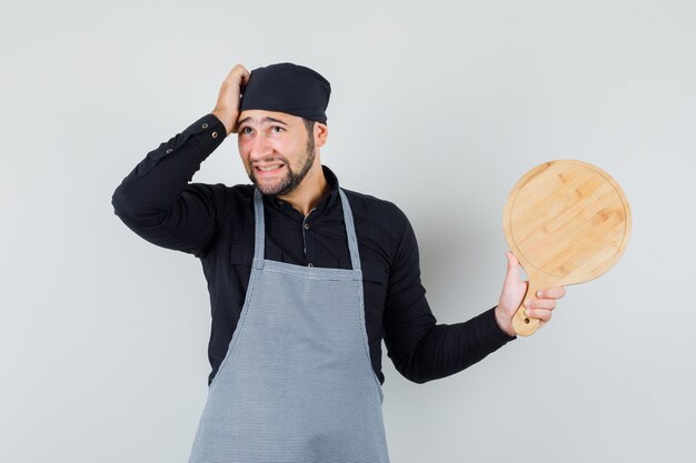 Young man holding cutting board while scratching head in shirt, apron and looking humble. front view.