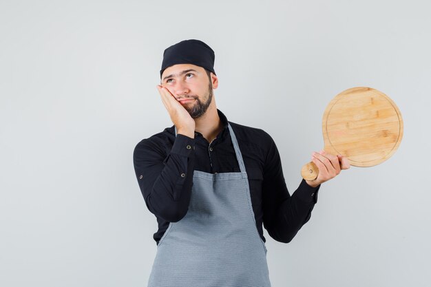 Young man holding cutting board with palm on cheek in shirt, apron and looking pensive. front view.