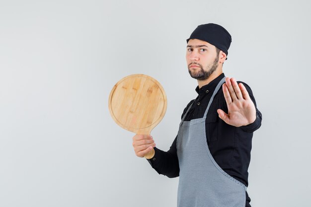 Young man holding cutting board with stop sign in shirt, apron and looking serious. front view.