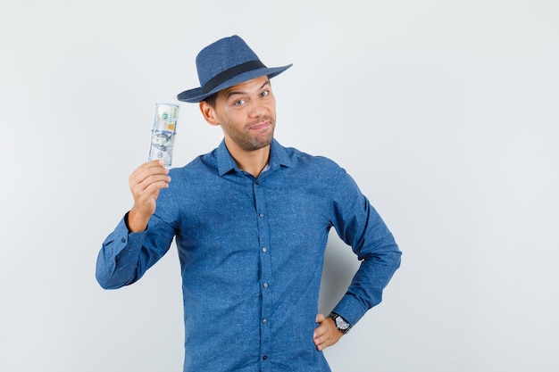 Young man holding dollar banknote in blue shirt, hat and looking merry. front view.