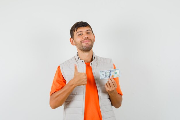 Молодой человек держит долларовую купюру, показывает большой палец вверх в футболке, куртке и выглядит веселым, вид спереди.