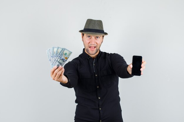Молодой человек держит долларовые купюры и смартфон в черной рубашке