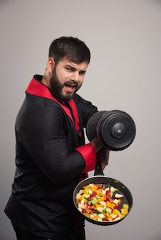 Young man holding a dumbbell and pan with vegetables.