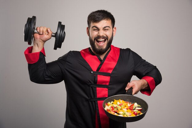 Young man holding a dumbbell and pan with vegetables.