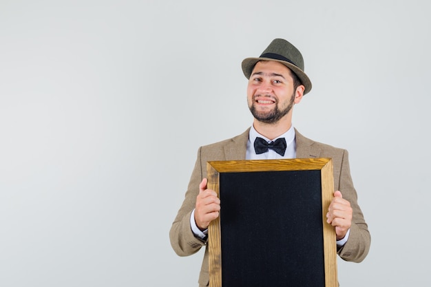Young man holding empty chalkboard in suit, hat and looking cheerful , front view.