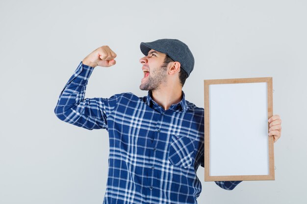 Young man holding empty frame in shirt, cap and looking happy , front view.