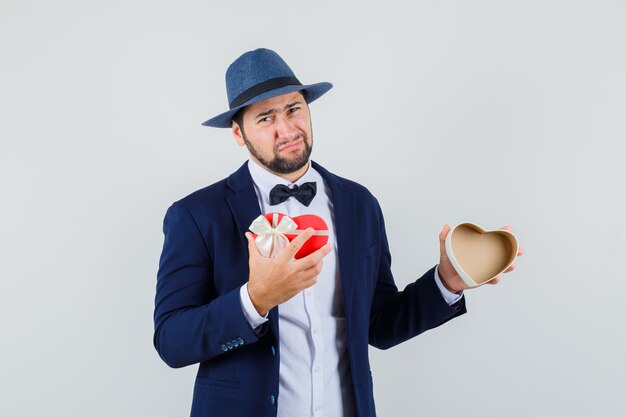 Young man holding empty present box in suit, hat and looking disappointed , front view.