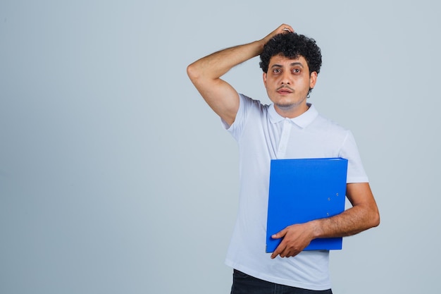 Young man holding file folder, holding hand on head in white t-shirt and jeans and looking pensive. front view.