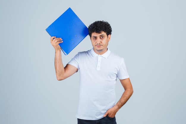 Молодой человек держит папку с файлами и держит одну руку в кармане в белой футболке и джинсах и выглядит серьезно. передний план.