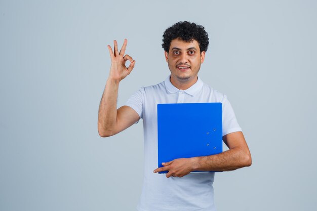 Young man holding file folder and showing ok sign in white t-shirt and jeans and looking happy , front view.